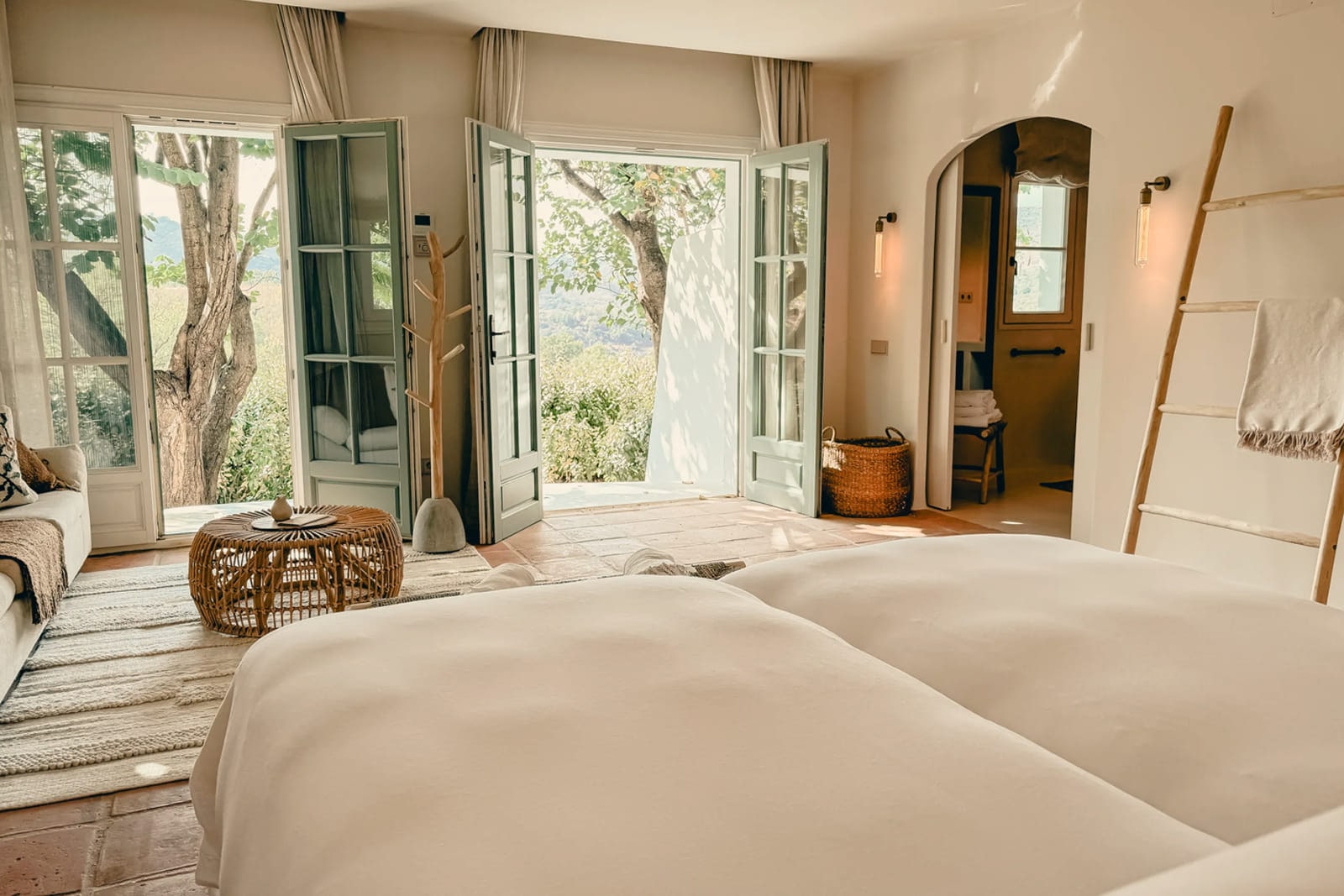 A bedroom at Viluz: white linen, a woven ladder, and french doors standing open to the garden.