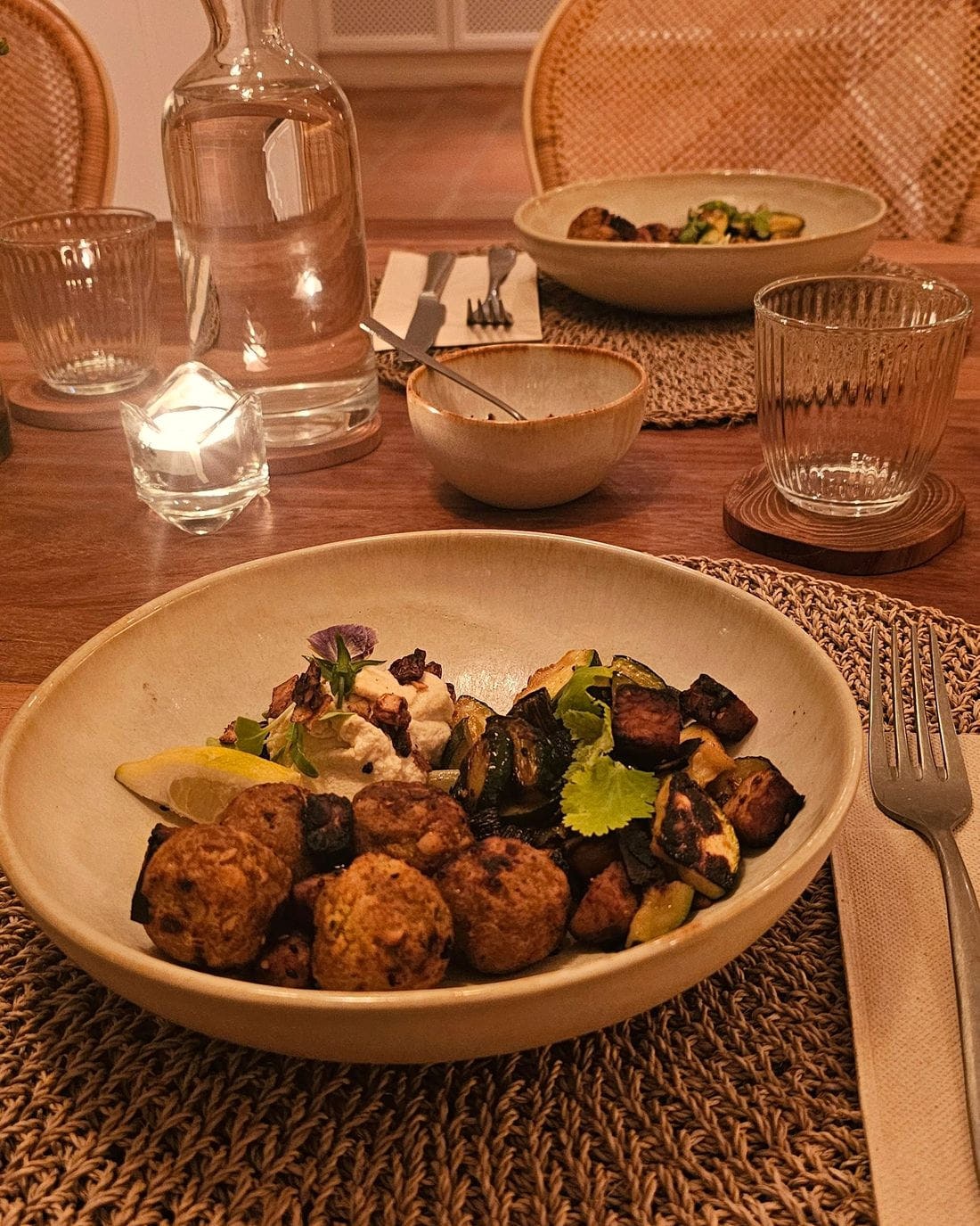 A plated dish of roasted vegetables in a ceramic bowl, lit by a single candle on a linen mat.