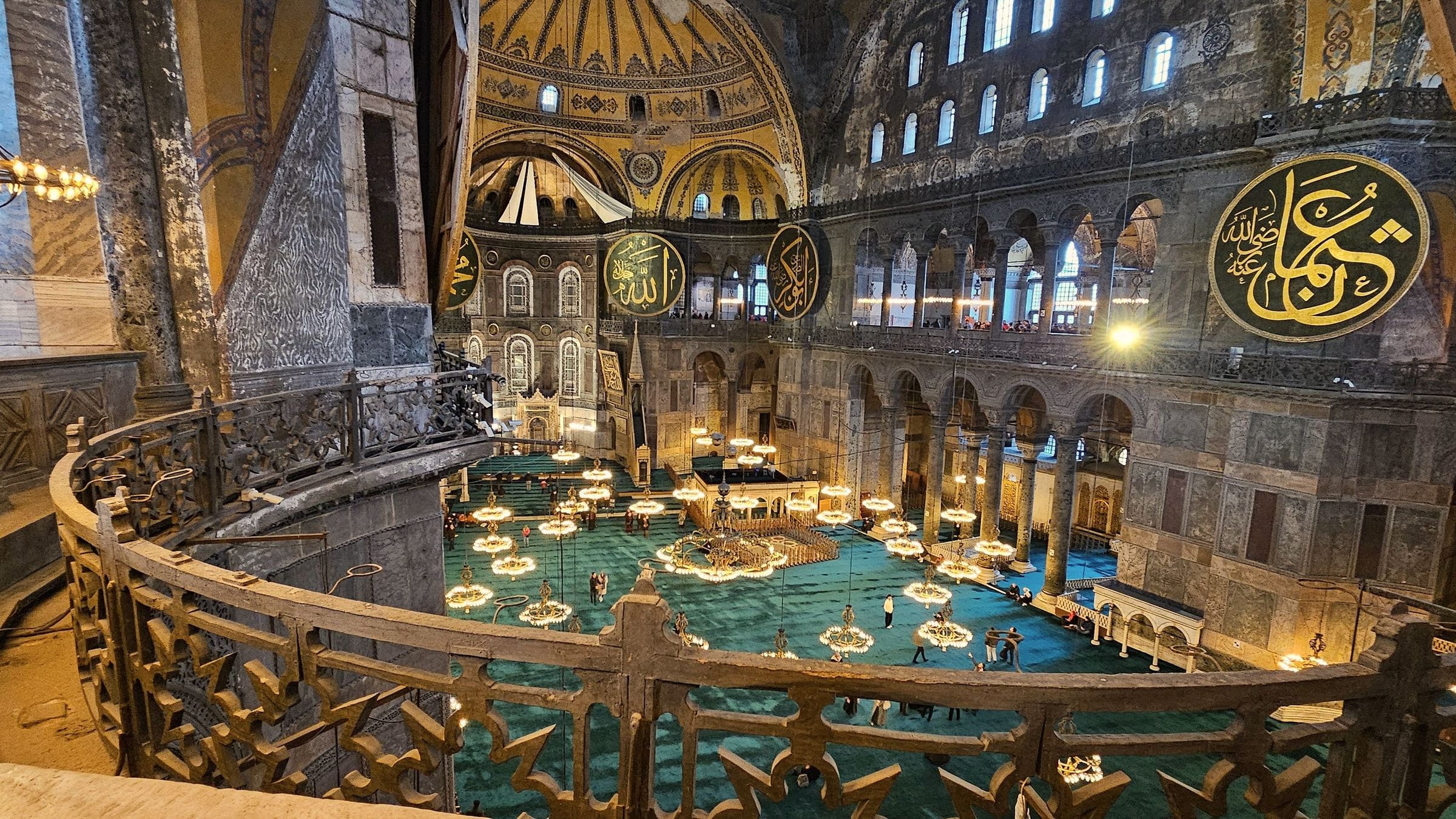 The interior of Hagia Sophia from the upper gallery: chandeliers over the nave, calligraphic medallions, and the great dome above.