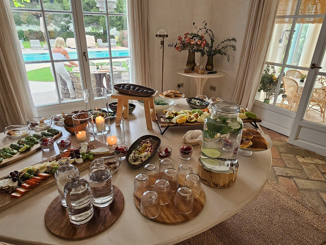 A long table laid with mezze, glasses and candles, tall windows open to the pool beyond.